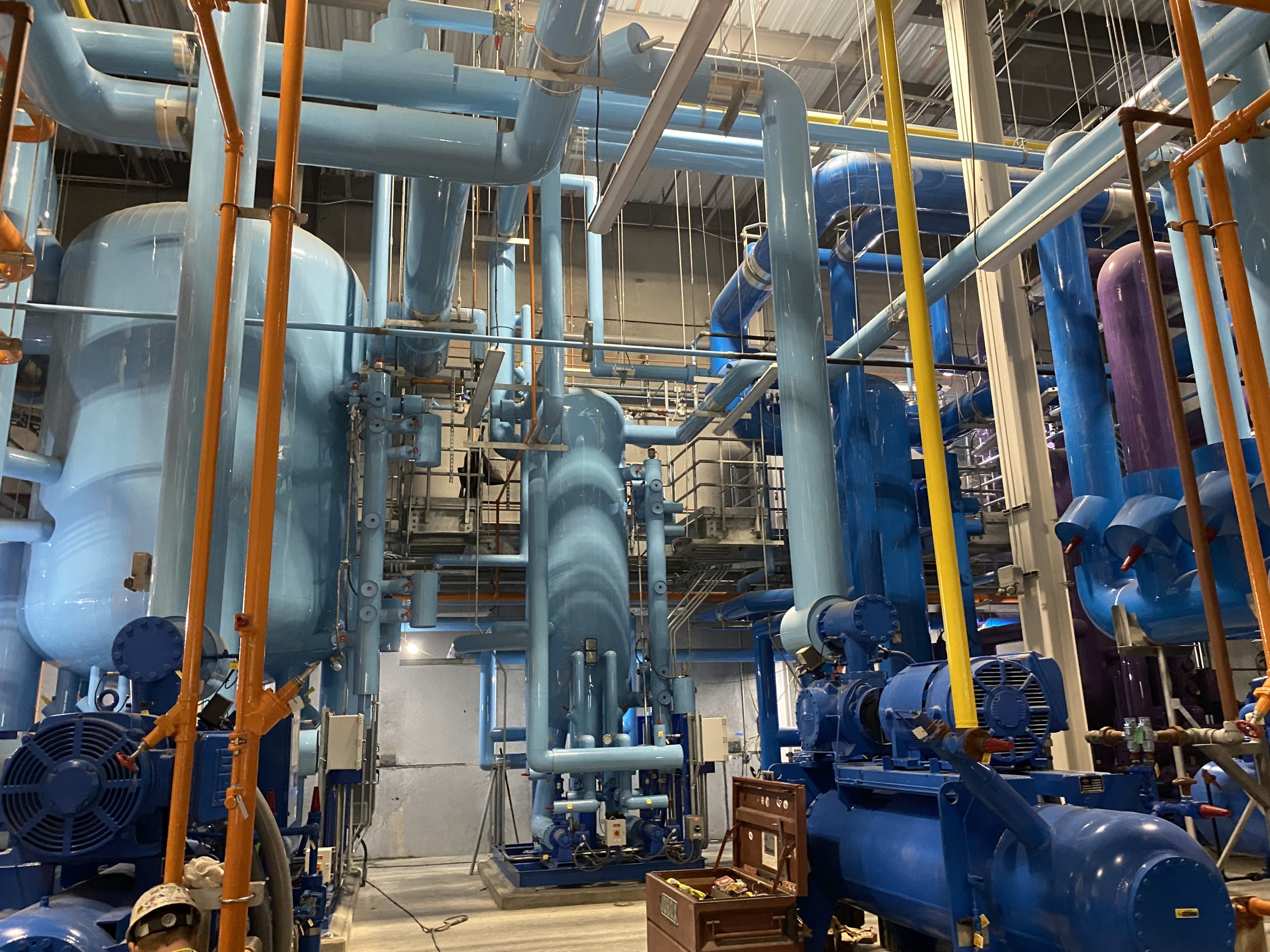 A complex industrial facility with interconnected blue piping and machinery, surrounded by orange pipes and other equipment, housed within a spacious interior.