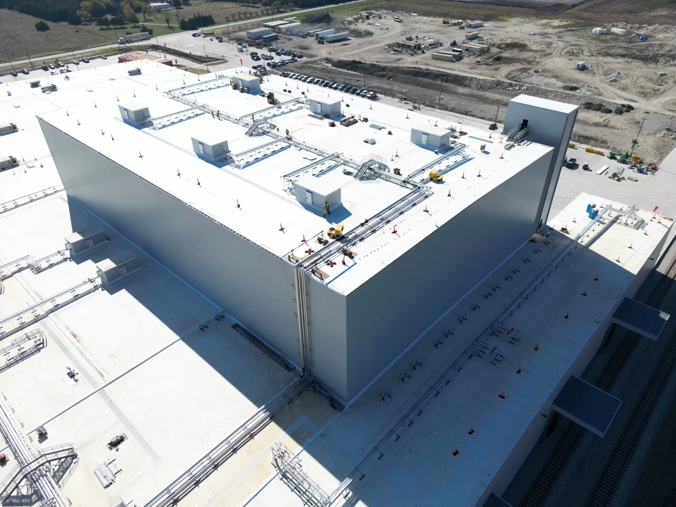 Aerial view of a large industrial building with extensive rooftop equipment. Surrounding area includes construction activity and earth-moving machinery.