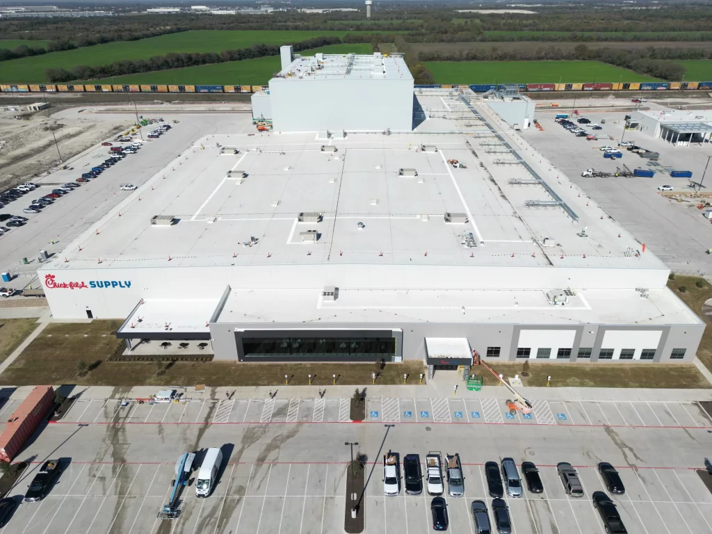 Aerial view of a large white industrial facility with "Supply" signage, surrounded by parking lots and loading docks. A train track runs parallel to the building, with fields in the background.