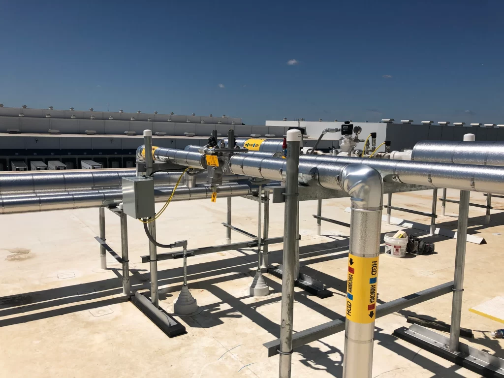 A network of insulated industrial pipes is installed on a rooftop. The pipes are secured with metallic supports and labeled with safety warnings. A clear sky is visible in the background.