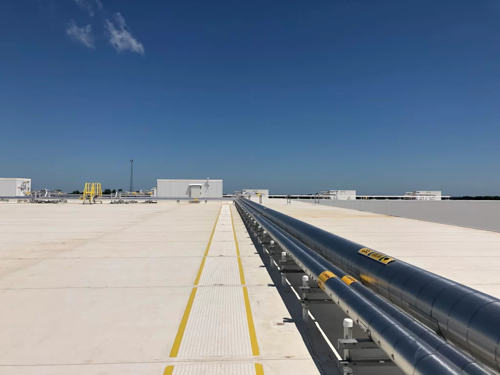 A long, metallic pipeline runs straight across a flat, light-colored surface under a clear blue sky. Industrial buildings and equipment are visible in the background.