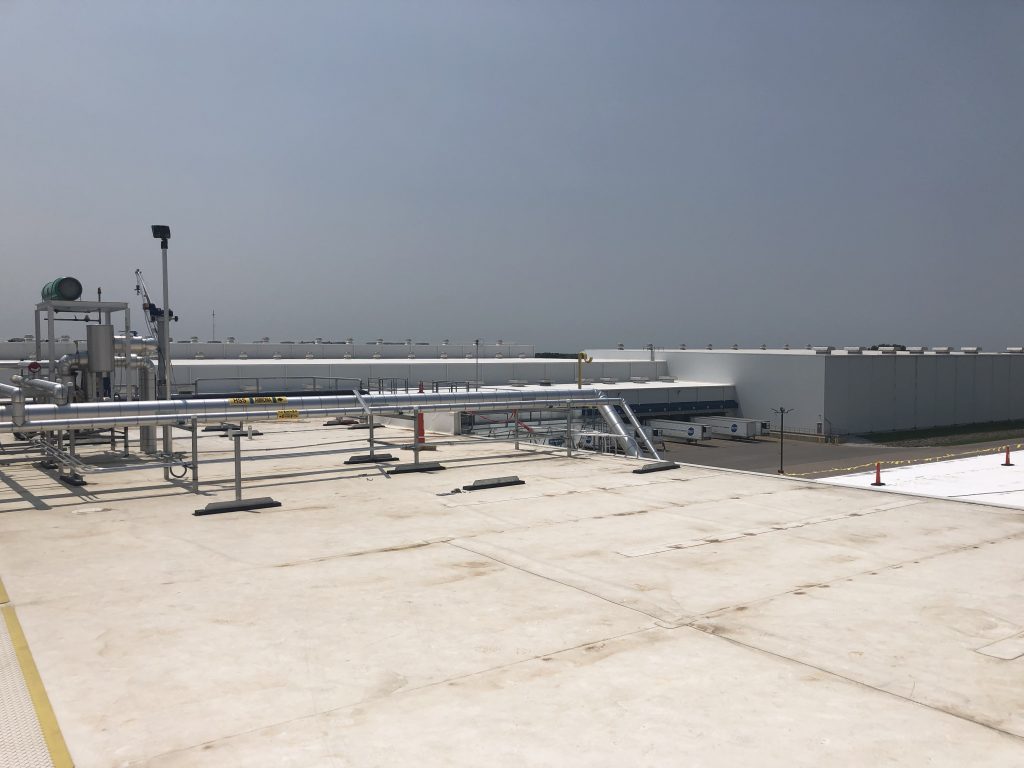 Flat rooftop with industrial pipes and equipment in the foreground, under a clear sky, and large white warehouse buildings visible in the background.