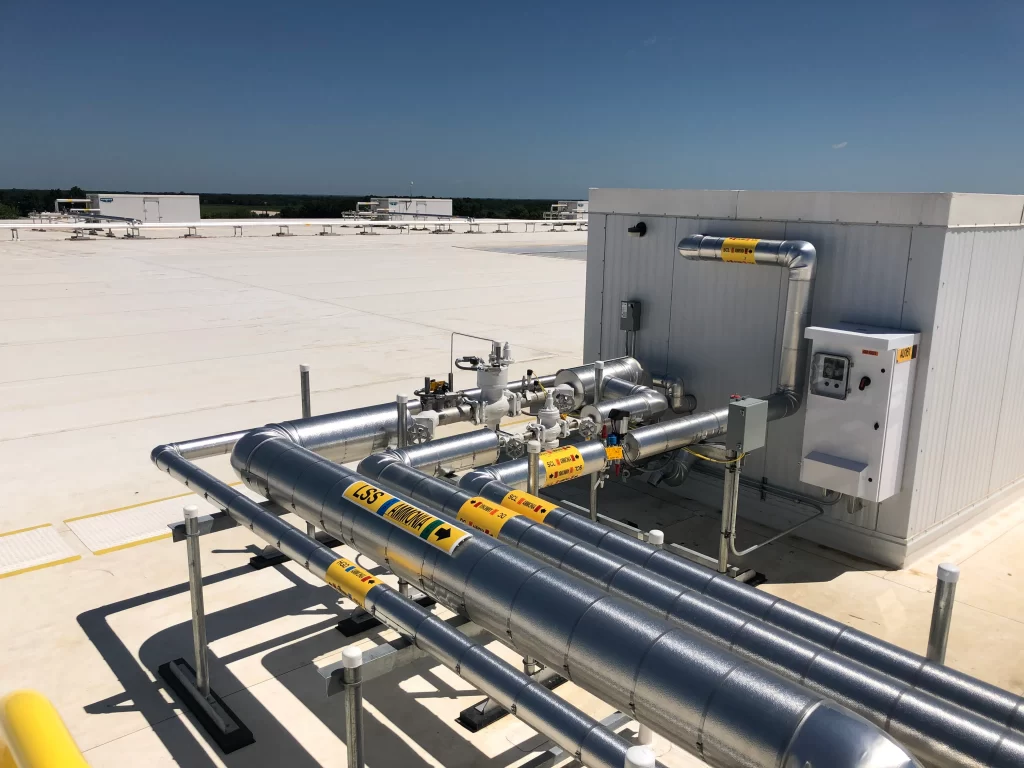 Industrial piping system on a rooftop with labeled metal pipes, a control panel, and a small white building under a clear blue sky.