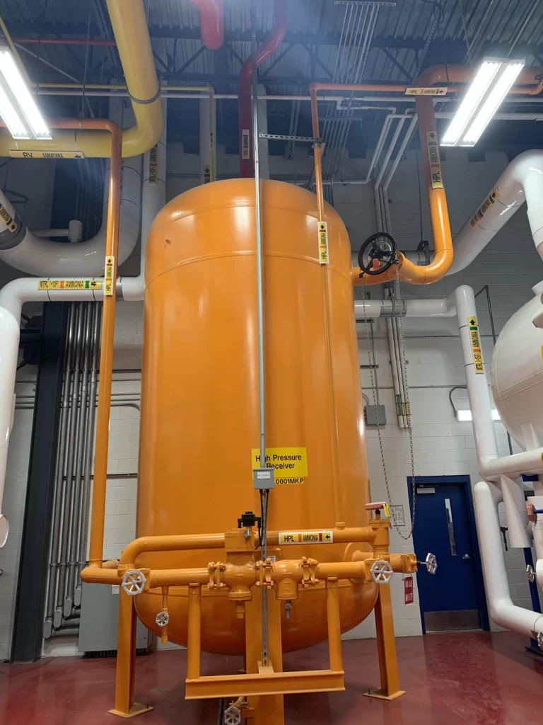 Large orange industrial pressure container with attached pipes, situated in a utility room with multiple overhead lights. Signs indicating hazardous areas and safety procedures are visible.