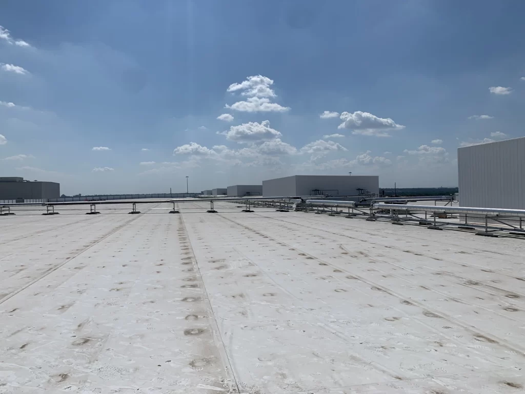 A flat, white rooftop under a clear, blue sky with scattered clouds. Several large white industrial buildings and metal railings are visible in the distance.