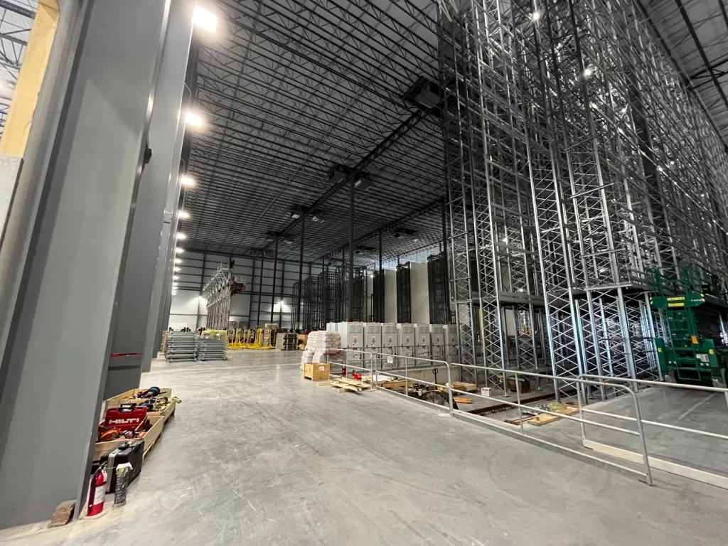 A large industrial warehouse with tall metal racks, assorted equipment, boxes, and construction materials scattered on the floor. High ceilings with exposed beams and lighting fixtures are visible.