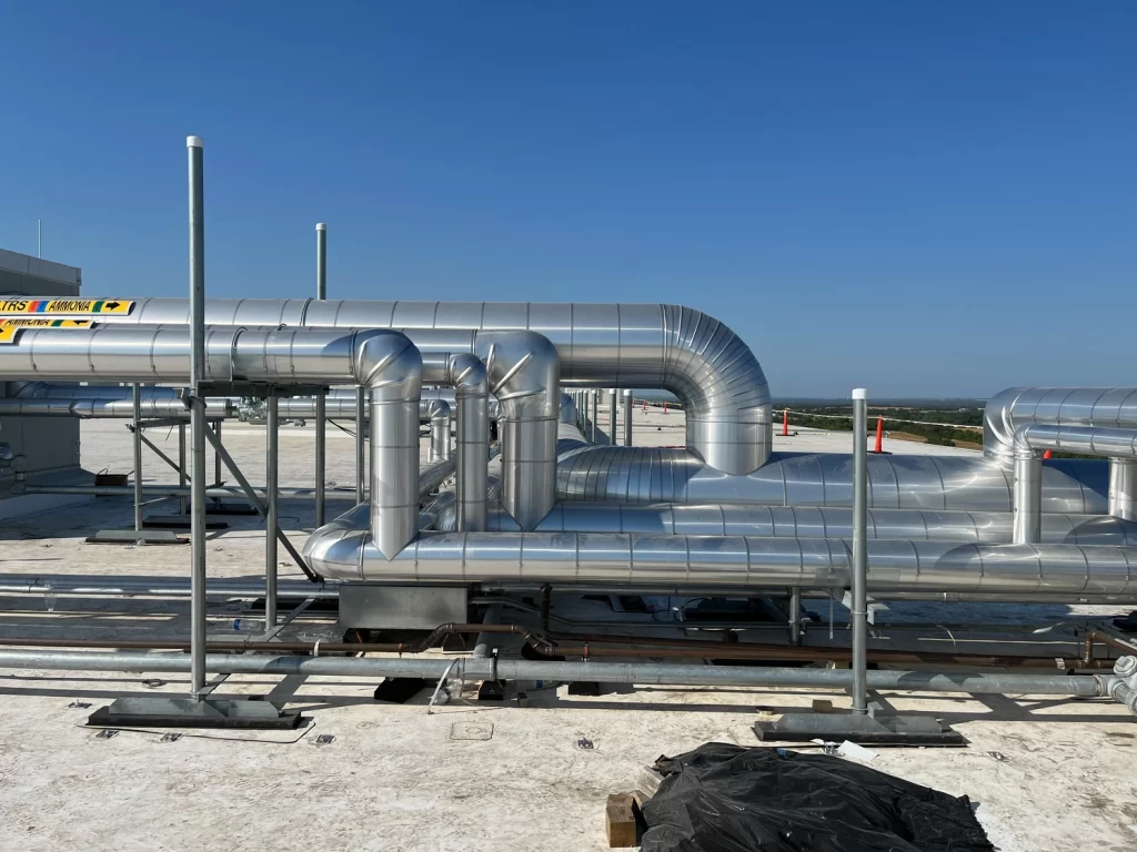 Metallic ductwork and ventilation pipes installed on a rooftop, with clear blue skies in the background. Construction materials and equipment are visible in the foreground.