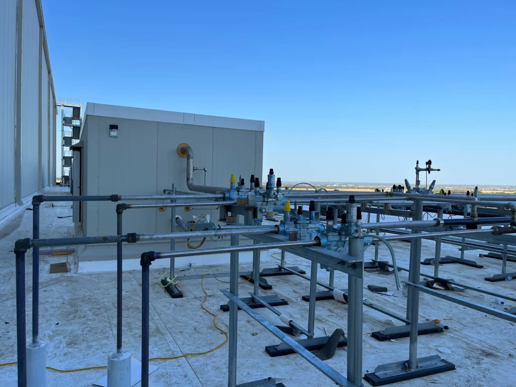 A rooftop view showing various mechanical and piping installations against a clear blue sky. Elevated equipment includes gas regulators, piping structures, and a ventilation system.