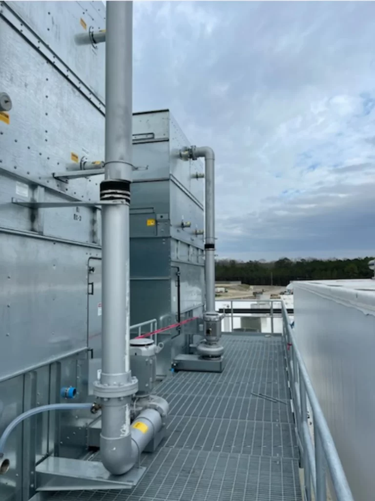 A rooftop industrial setup with large metal pipes and ventilation ducts against a cloudy sky background. Rails line a grated walkway beside the equipment.