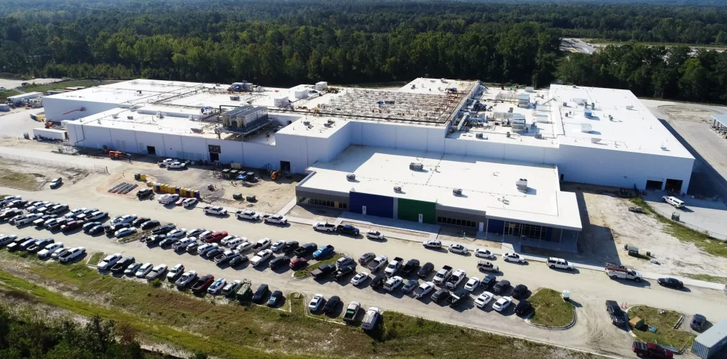 Aerial view of a large industrial building under construction, surrounded by a parking lot filled with numerous cars and trucks, set against a backdrop of dense trees.