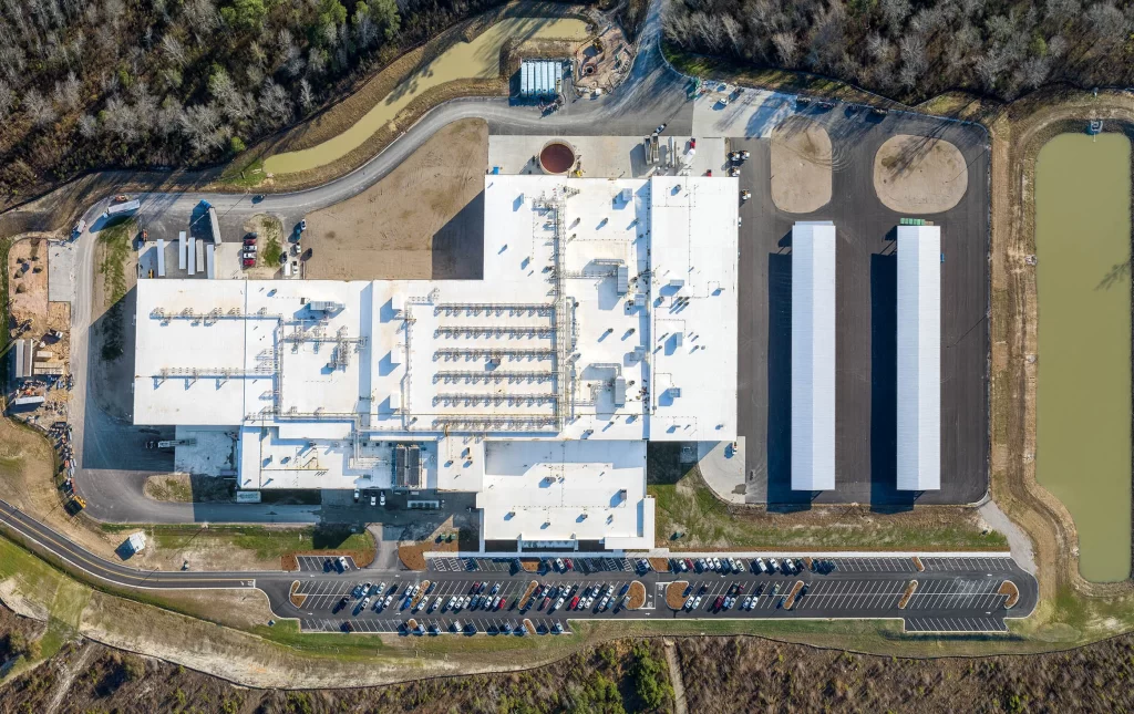 Aerial view of a large industrial facility with several buildings, parking area, ponds, and surrounding forested area.