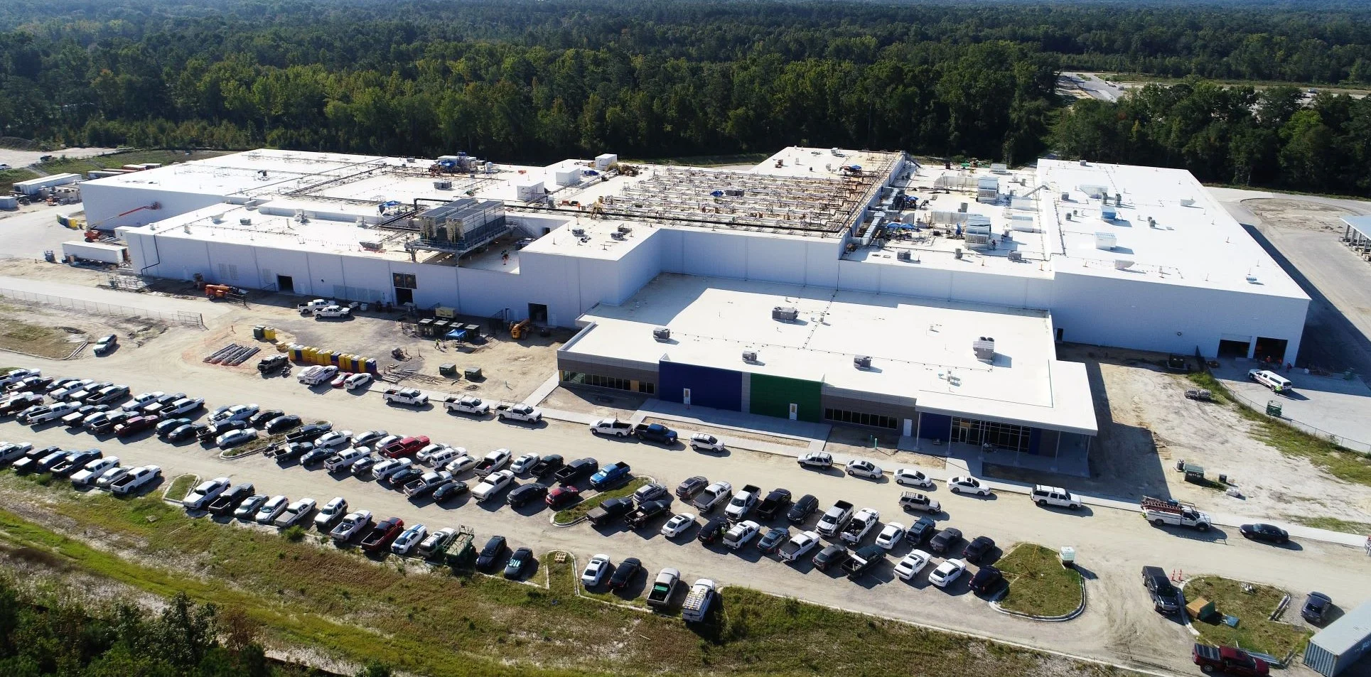 Aerial view of a large industrial building under construction, surrounded by a parking lot filled with numerous cars and trucks, set against a backdrop of dense trees.