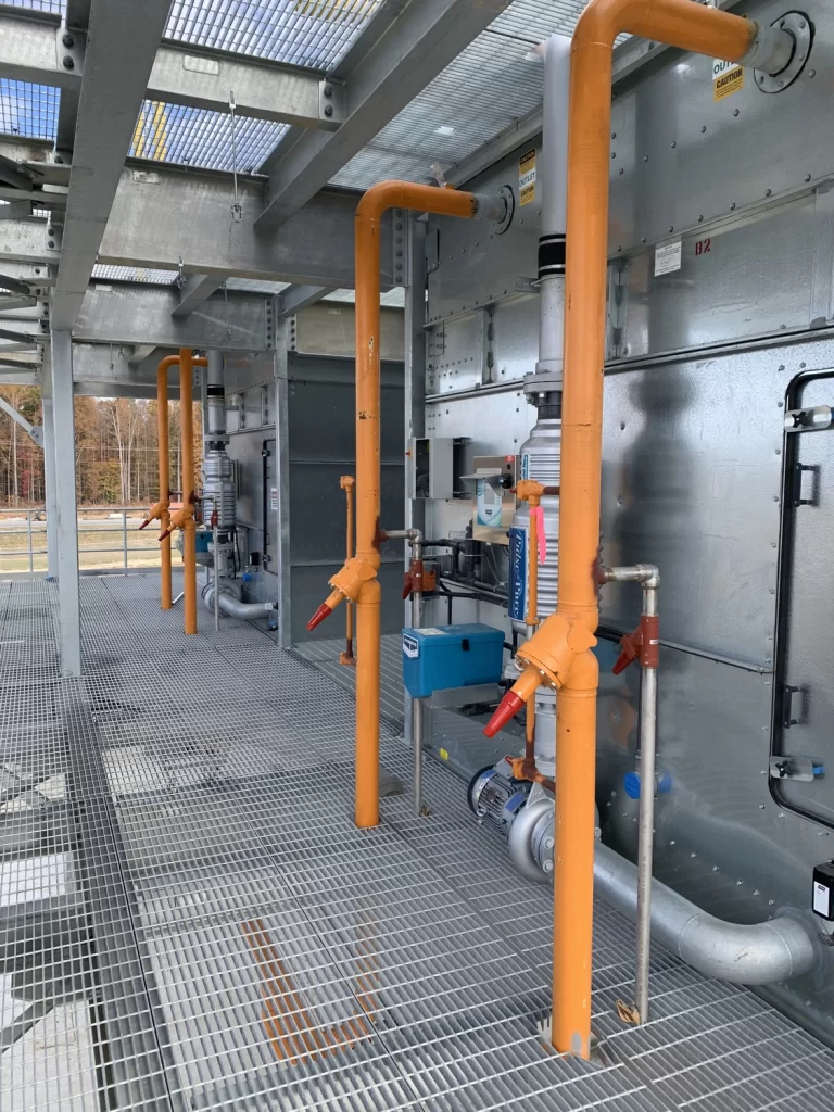 Industrial piping system with orange pipes, shut-off valves, and metal grating floor within an indoor facility.