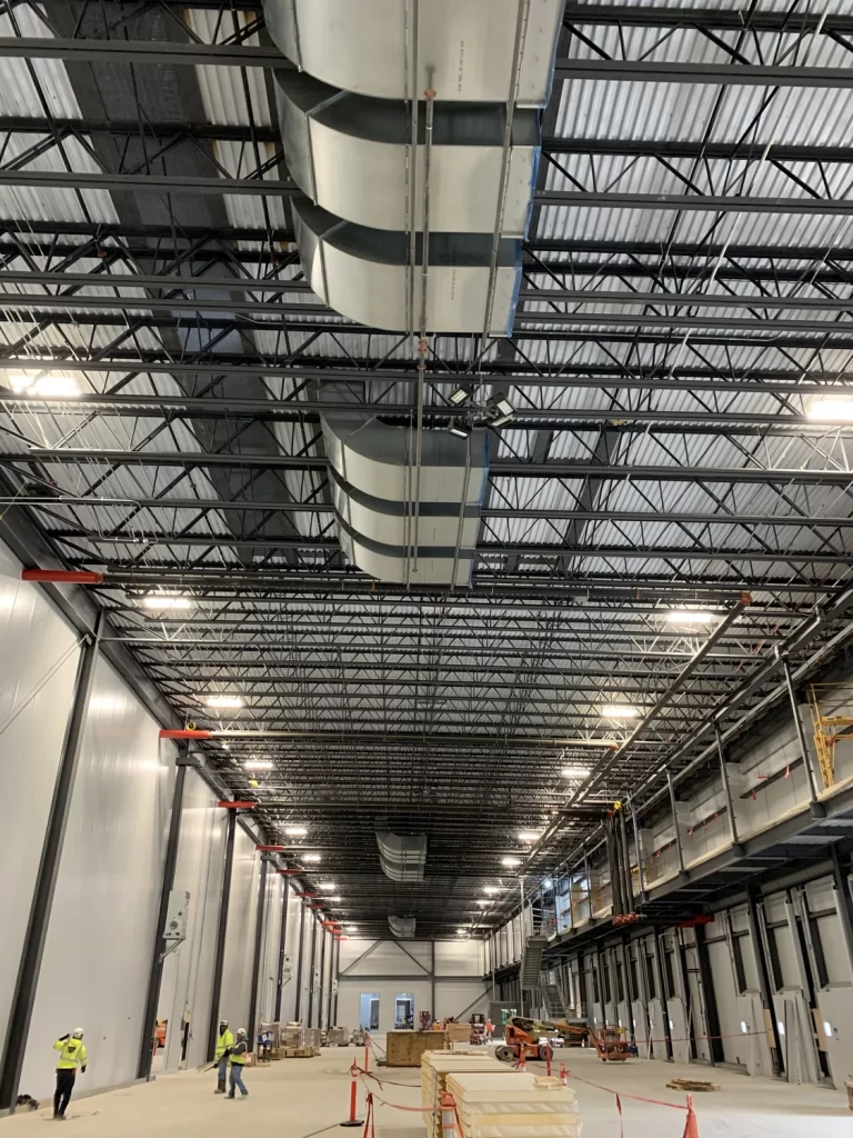 Large industrial warehouse under construction with high ceilings, exposed ductwork, and steel beams. Workers, construction materials, and equipment are visible on the floor.