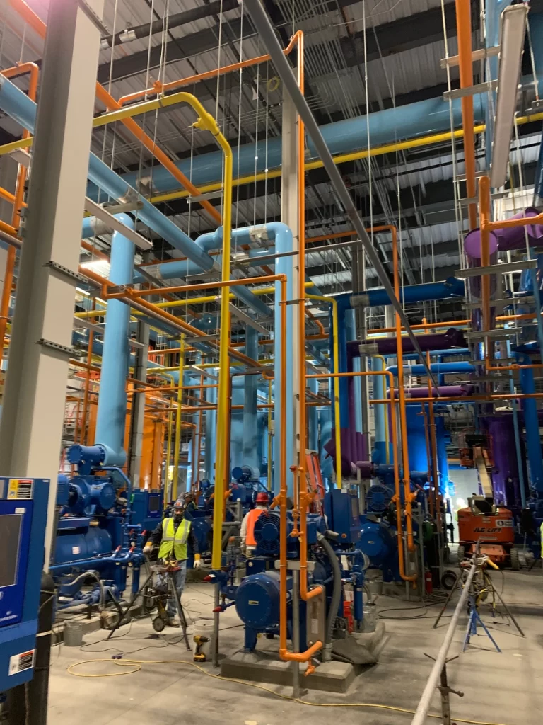 An industrial facility showing a complex arrangement of colorful pipes and machinery with workers in high-visibility clothing inspecting the equipment.