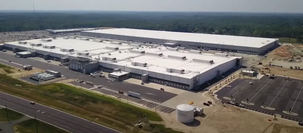 Aerial view of a large industrial warehouse complex surrounded by parking lots and roads, set in a rural area with trees in the background.