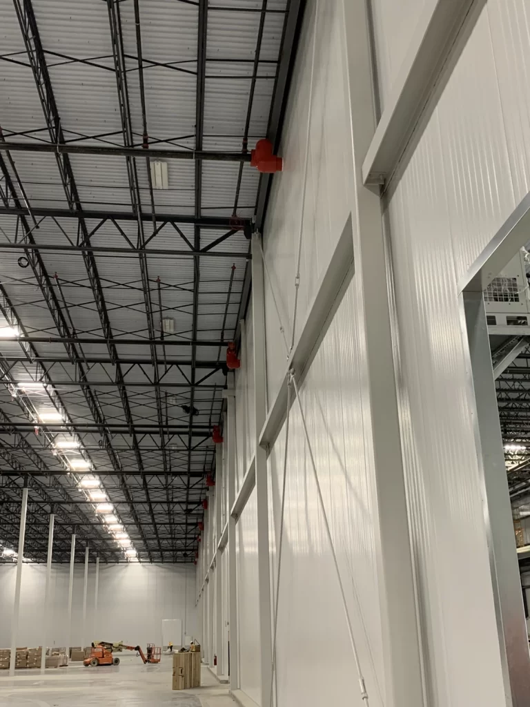 Interior of a large industrial warehouse with high ceilings, exposed steel beams, and white walls. Scattered pallets and equipment are on the floor, and some machinery is visible in the background.