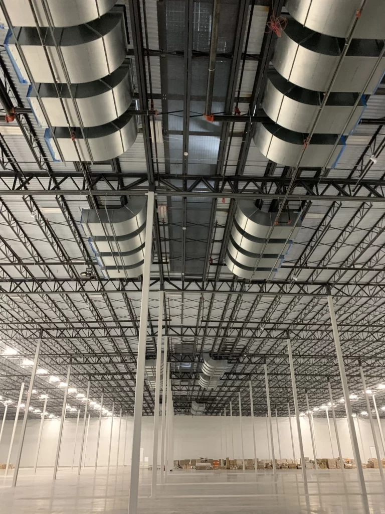 A spacious industrial warehouse interior with a high ceiling, metal beams, ductwork, and rows of support columns. The floor is bare, and there are a few boxes along the far wall.