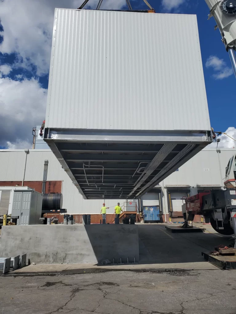 A large, white industrial unit being hoisted by a crane outdoors, with two workers in yellow vests standing nearby. White building and blue sky with clouds in the background.