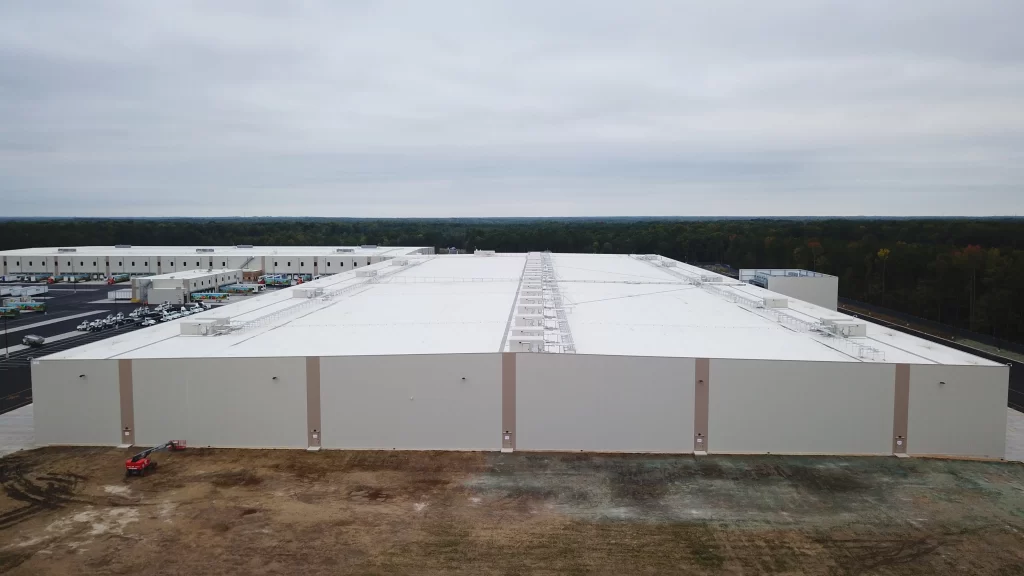 Aerial view of a large, white-roofed industrial warehouse with adjacent smaller buildings and a sparse parking area under a cloudy sky.