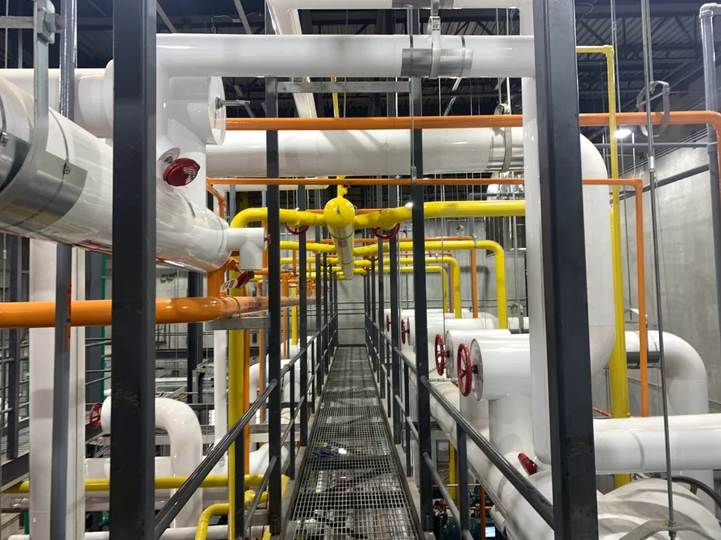 A network of industrial pipes and valves of various colors, primarily white, yellow, and orange, is connected within a facility, with a metal walkway running through the center.