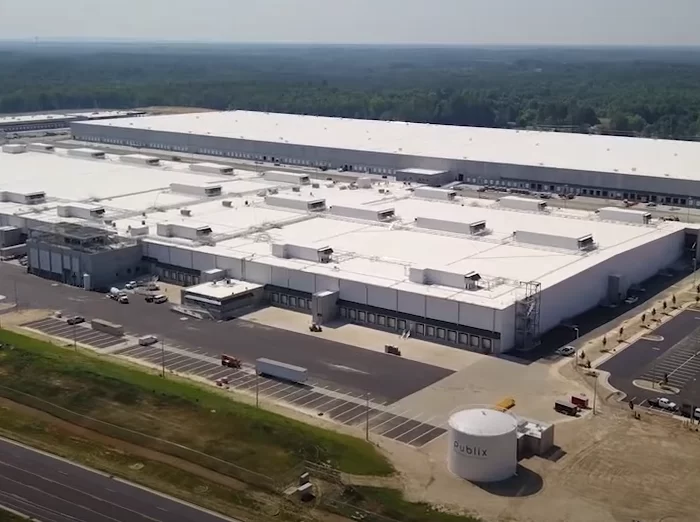 Aerial view of a large, white roofed industrial facility surrounded by parking lots and a few vehicles, located near a road with forest in the background.