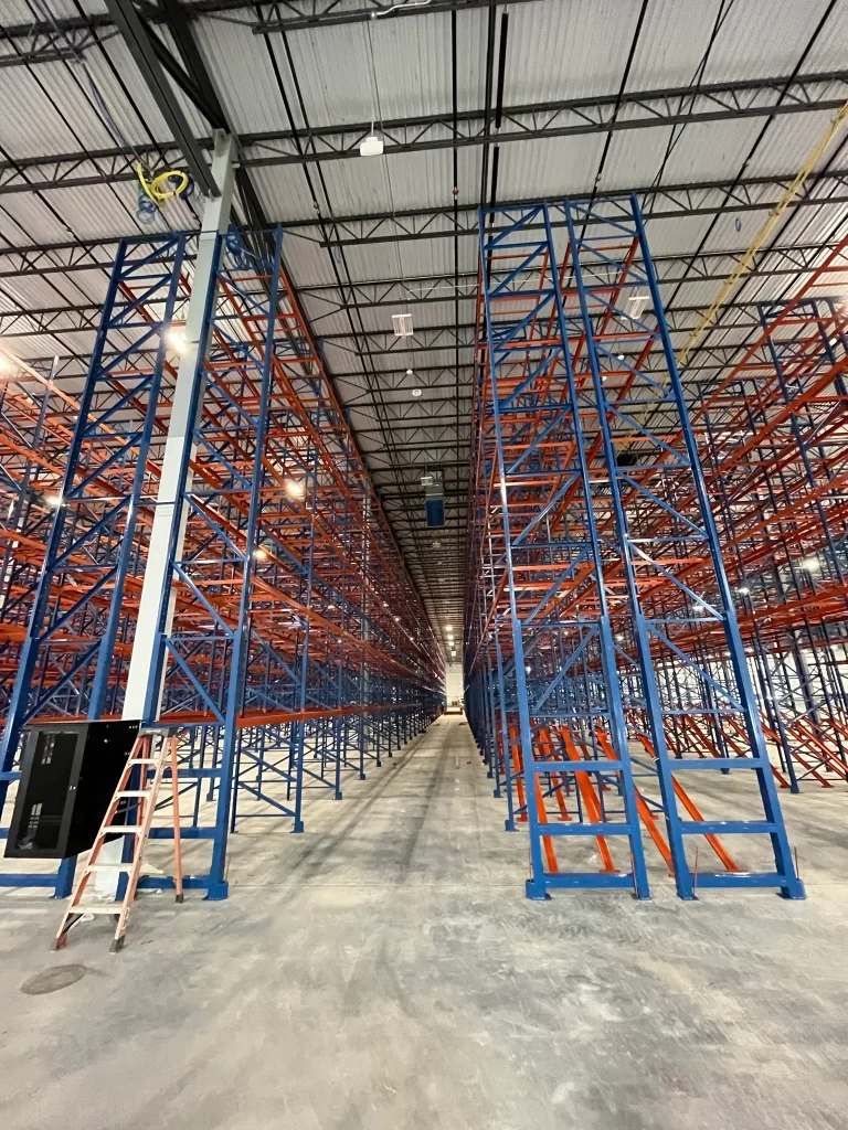 Empty warehouse interior with tall blue and orange metal shelving units, a ladder to the left, and concrete flooring. Ceiling lights illuminate the space.