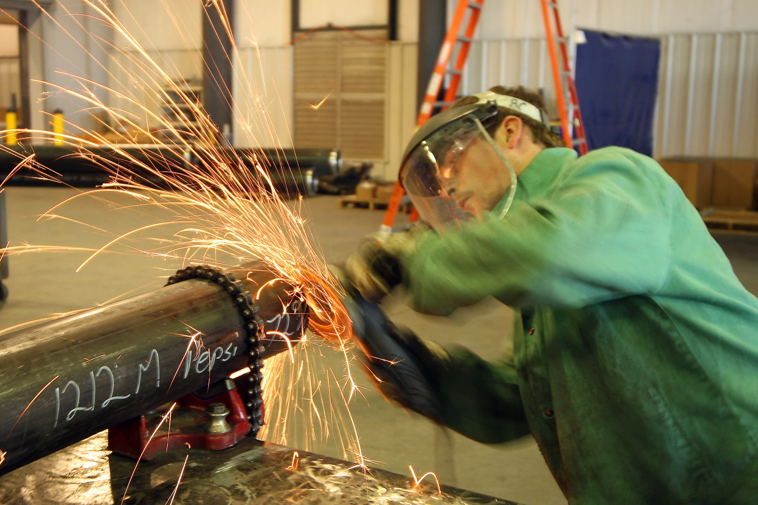 A worker wearing protective gear uses a cutting tool, producing sparks, on a metal pipe inside a warehouse with industrial equipment and ladders in the background.