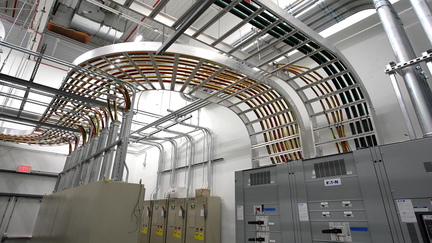 Metal cable trays with numerous colorful cables are mounted overhead in an industrial room with electrical equipment, panels, and labeled boxes against white walls.