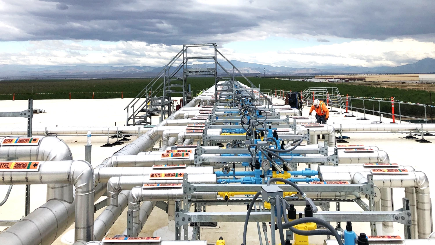 An industrial worker in protective gear inspects a series of interconnected pipes and valves on a large outdoor platform under a cloudy sky.