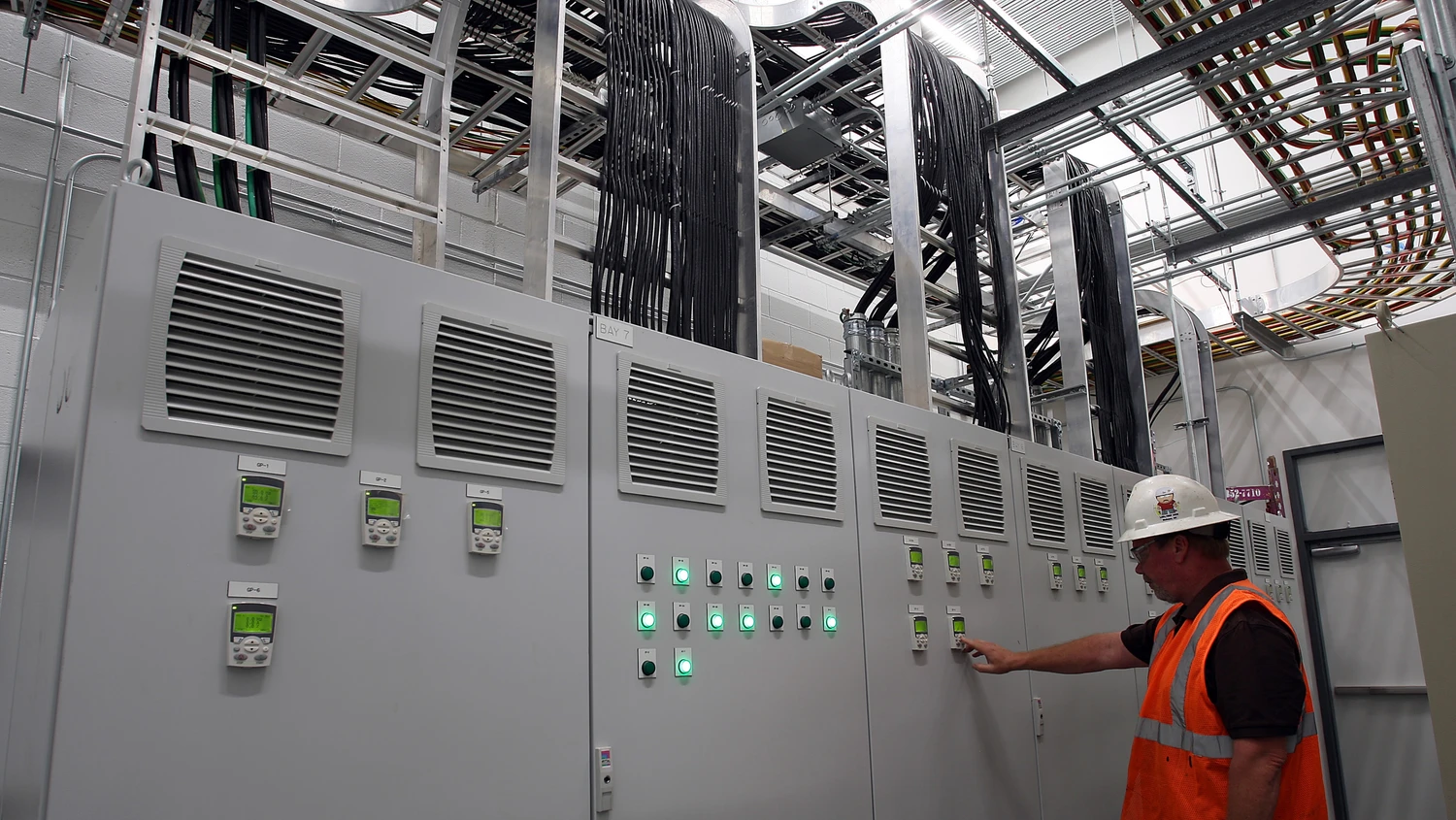 A worker in an orange safety vest and white helmet operates a control panel with illuminated green lights in an industrial setting with numerous cables overhead.