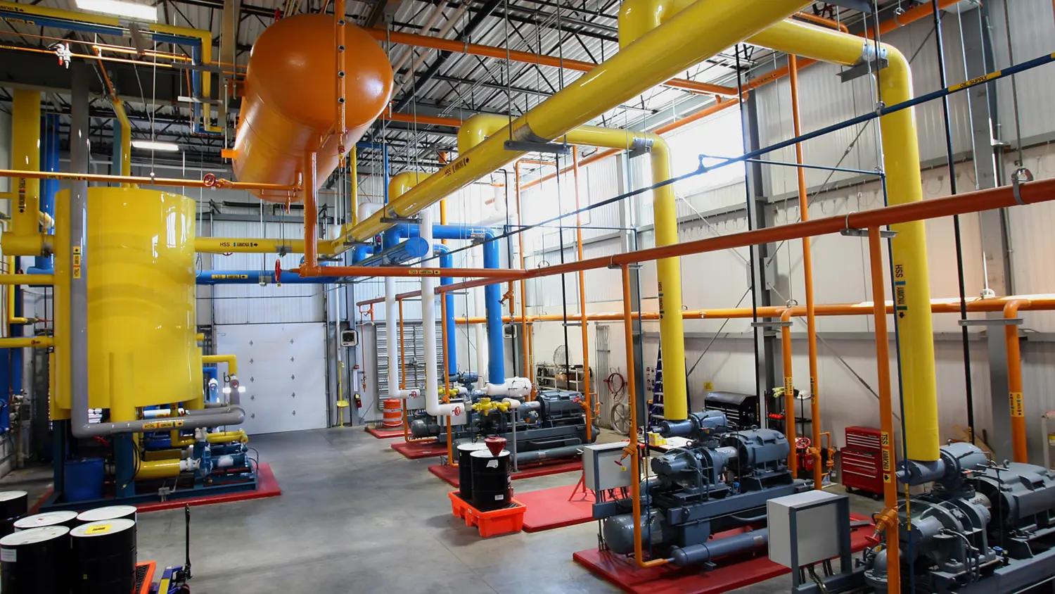 An industrial facility featuring bright yellow pipes, large tanks, and various machinery situated on a polished concrete floor, with a high ceiling and ample overhead lighting.