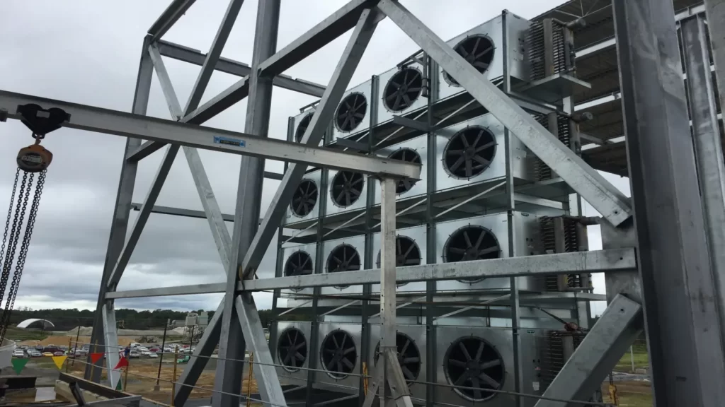 Tall metal structure with multiple large industrial fans arranged in a grid pattern, surrounded by steel framework. Construction site and cloudy sky in the background.