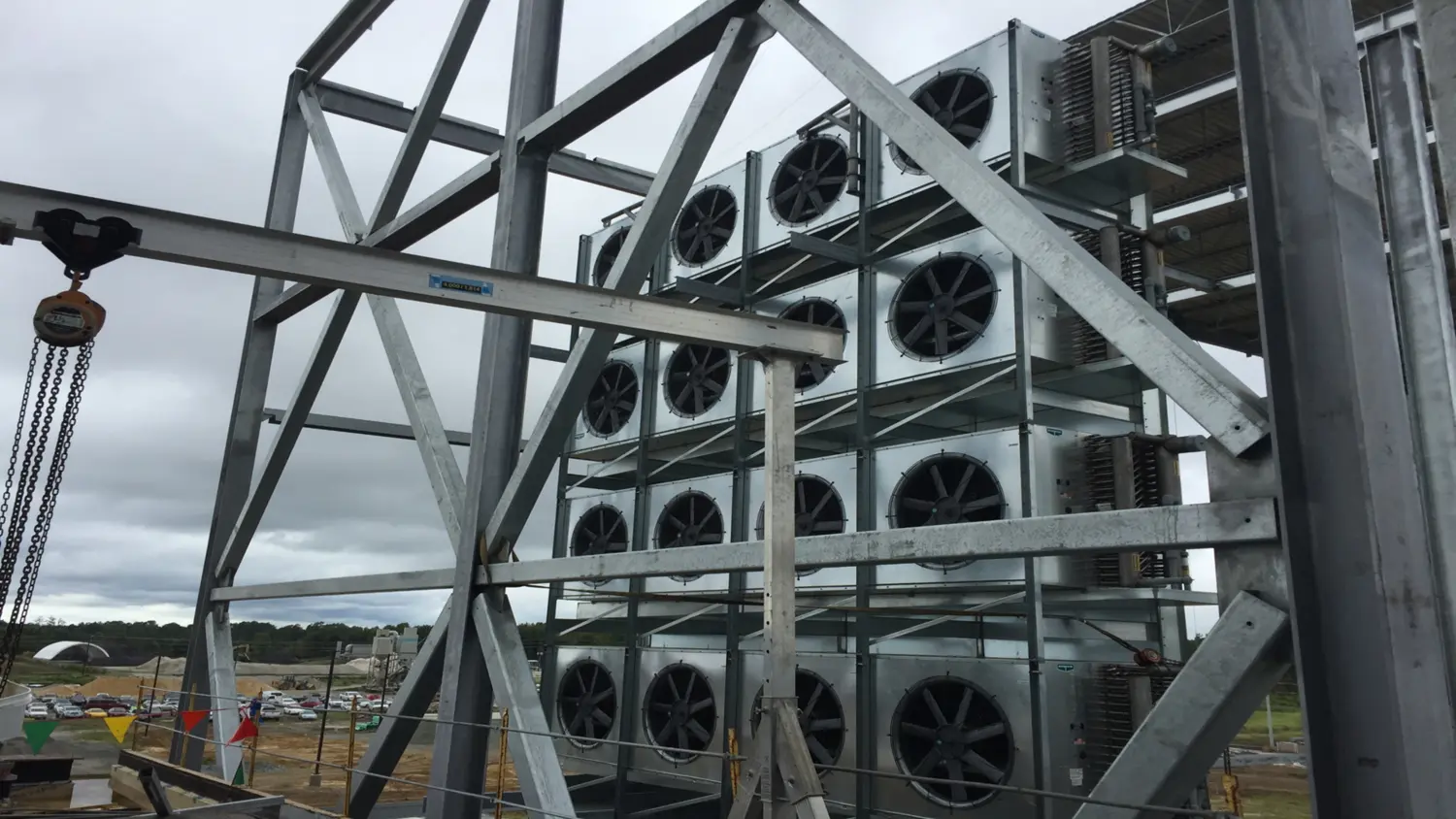 Tall metal structure with multiple large industrial fans arranged in a grid pattern, surrounded by steel framework. Construction site and cloudy sky in the background.