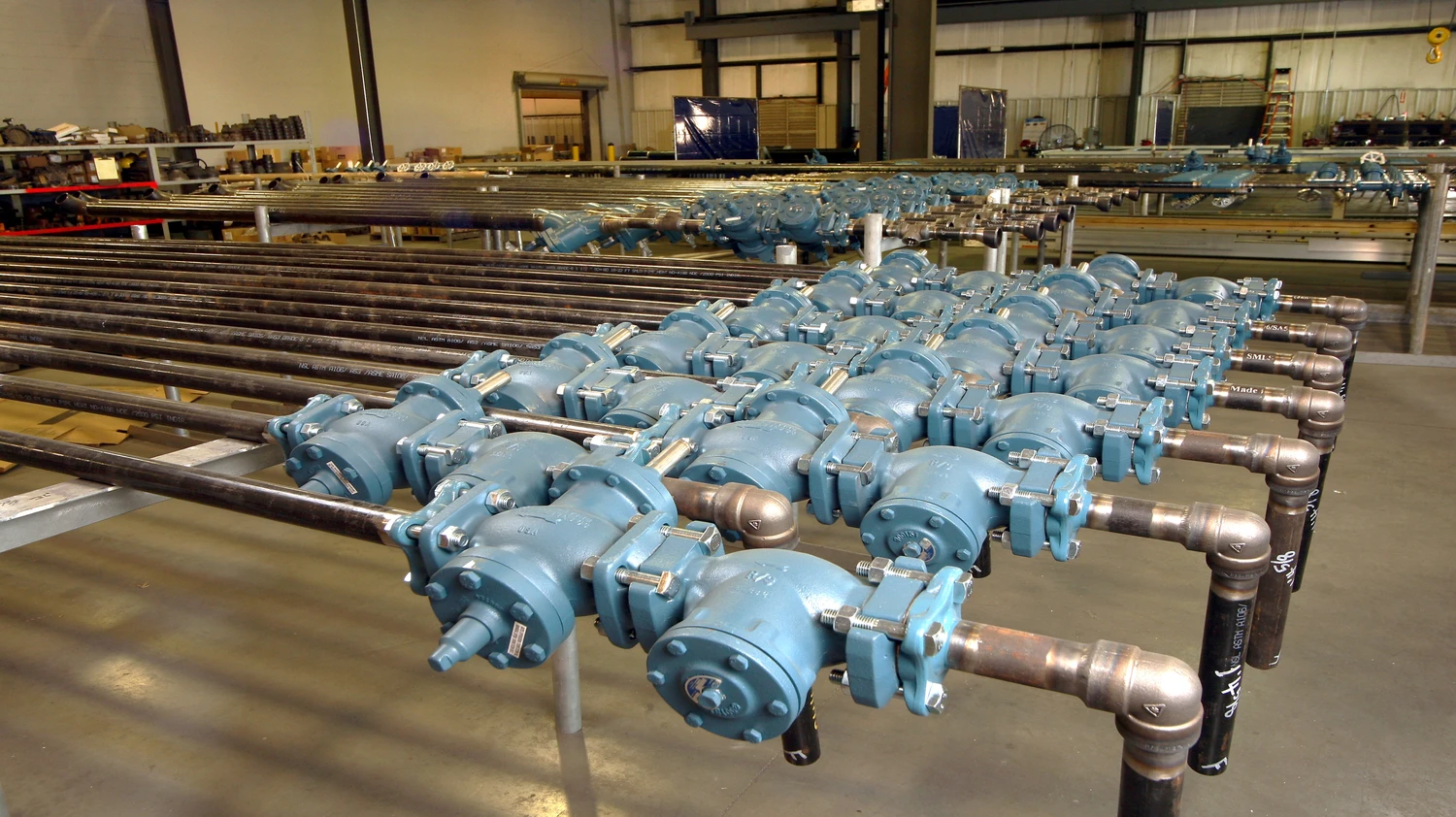 Multiple blue industrial pumps are aligned in rows inside a large warehouse. Various pipes and components are visible, with a concrete floor and industrial shelving in the background.