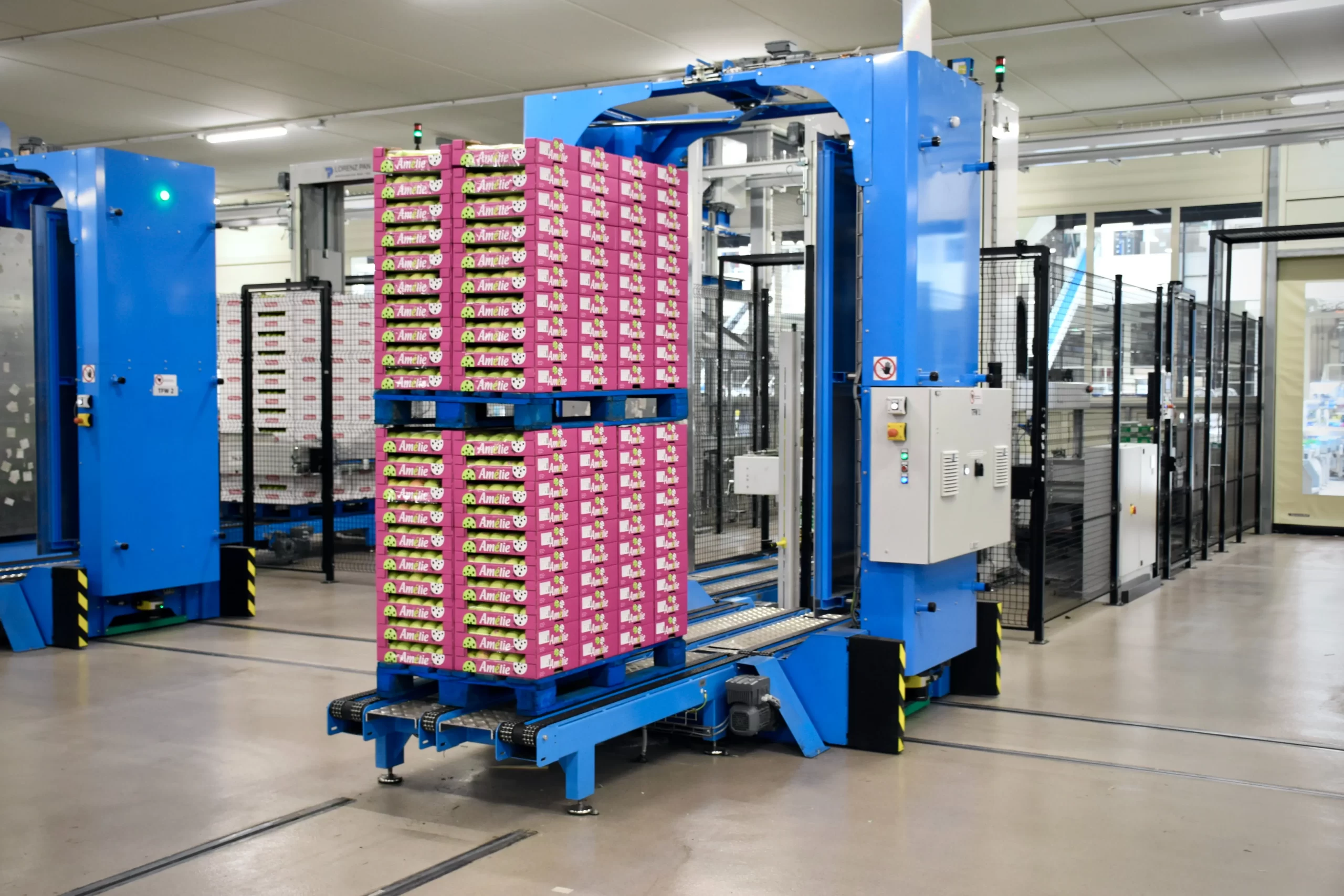 Automation machine with blue framework handling and moving a large stack of pink boxes filled with wrapped products in a factory setting.