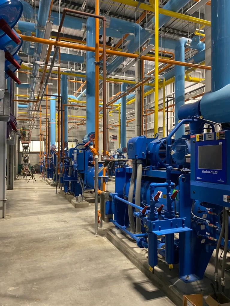 A large industrial facility featuring blue machinery and pipes, with orange and yellow piping overhead.