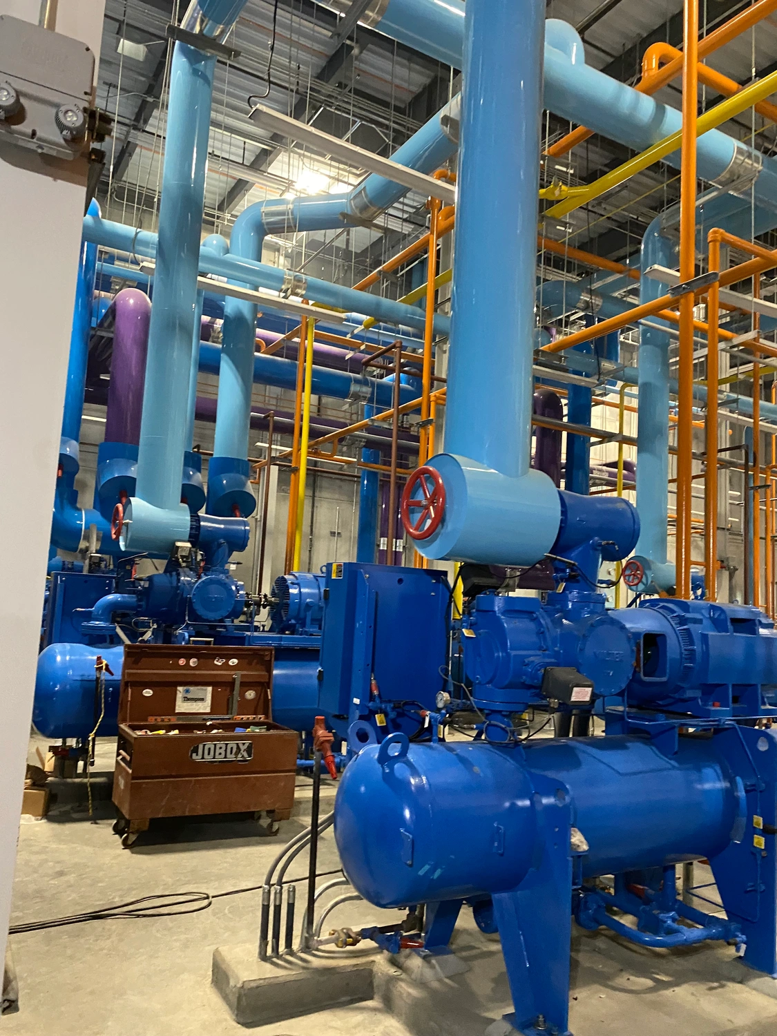 A room filled with industrial machinery and large blue pipes, with various valves and interconnected metal structures visible.