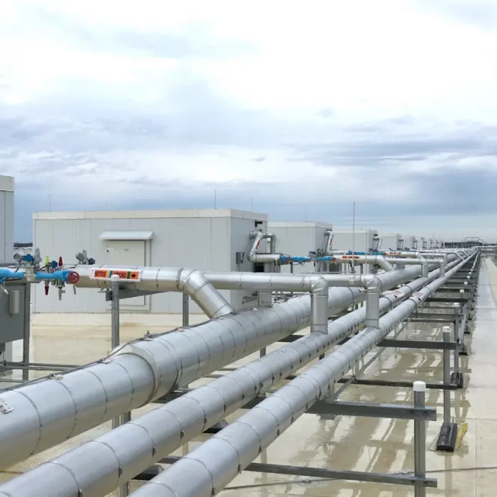 A row of metallic pipes and rooftop structures are seen against a cloudy sky in an industrial setting.