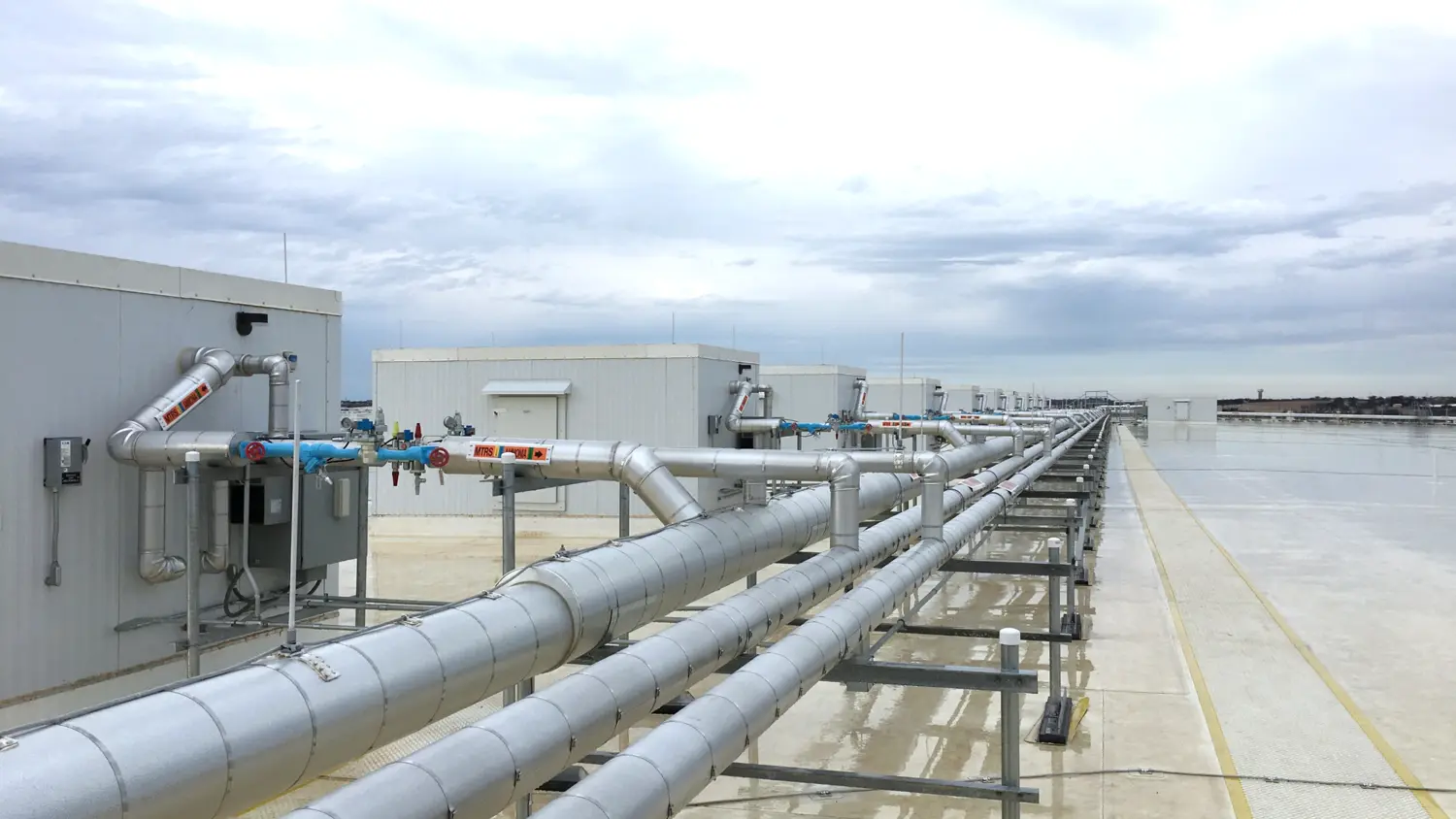 A row of metallic pipes and rooftop structures are seen against a cloudy sky in an industrial setting.