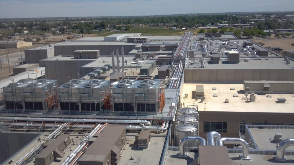 Aerial view of an industrial facility with multiple large cooling towers, pipelines, and rectangular buildings. The surroundings include open fields, residential areas, and distant greenery.