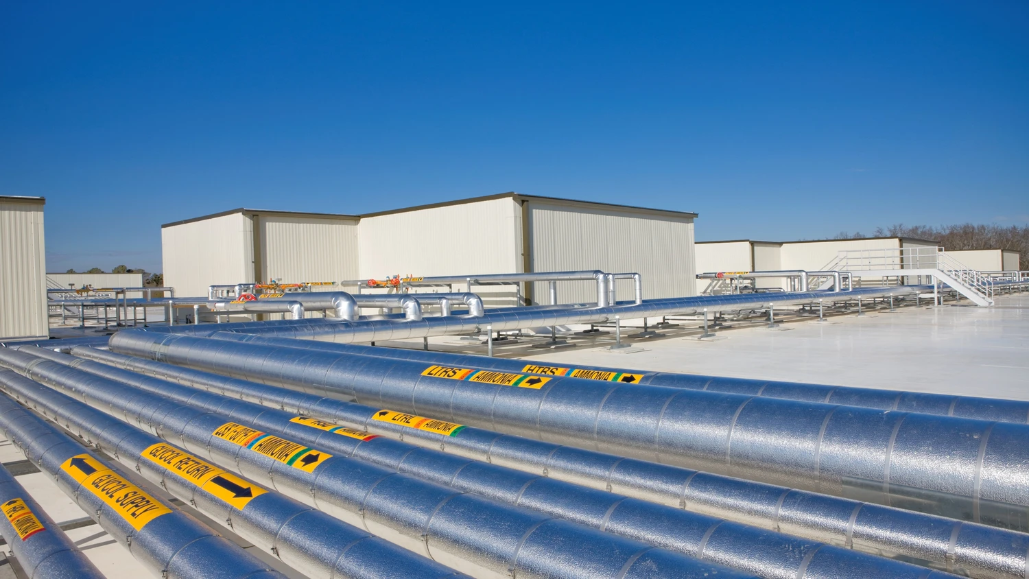 Multiple large, silver pipelines with safety labels are laid out on a reflective surface, leading to white industrial buildings under a clear, blue sky.