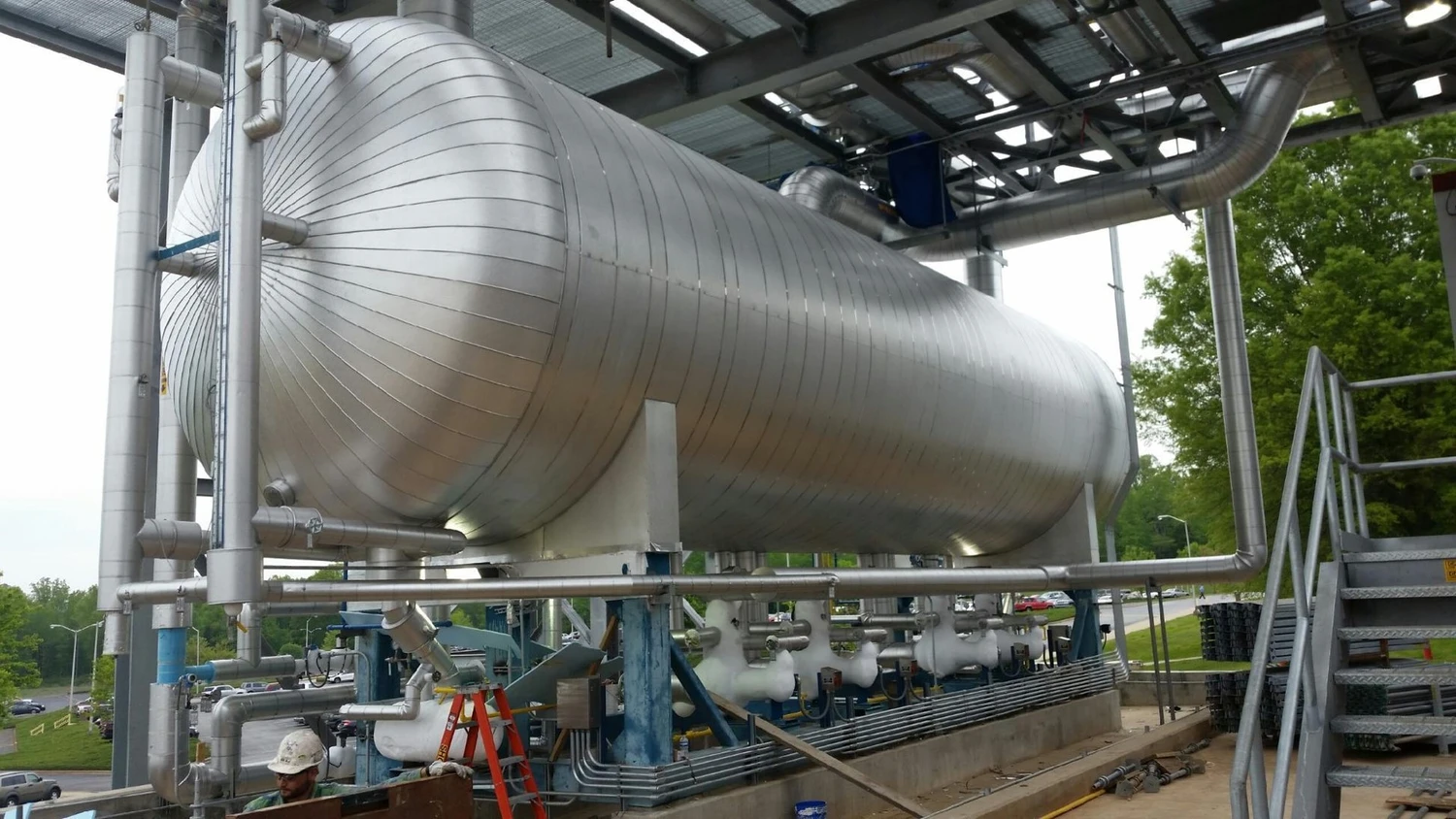 A large industrial storage tank made of metal is supported by white metal beams and connected to various pipes and valves. A worker in a hard hat is positioned near the base of the tank.