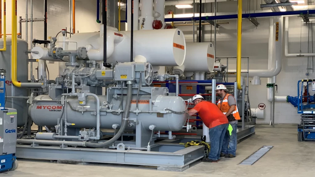 Two workers in safety gear inspect large industrial equipment in a brightly lit facility.