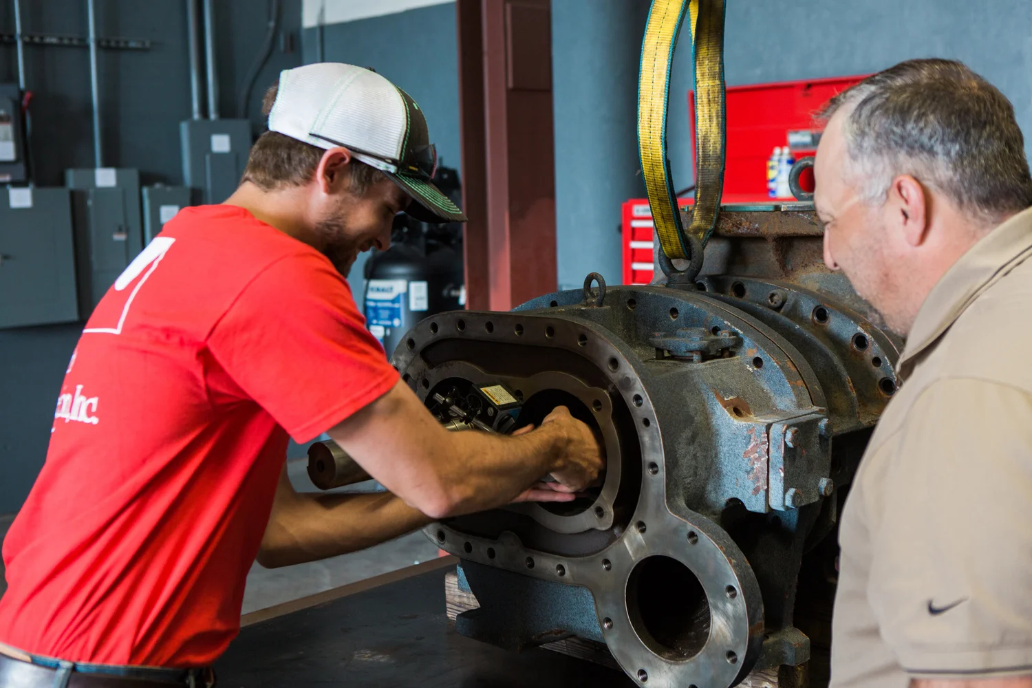 Two men inspect and work on a large, industrial metal component in a workshop with various tools and equipment in the background.