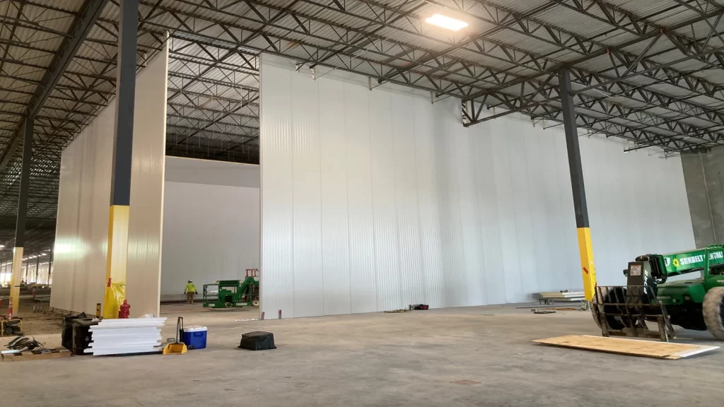 Large industrial warehouse interior with high white partition walls under construction. Scattered equipment and tools are visible on the concrete floor. Ceiling features metal beams and lighting.