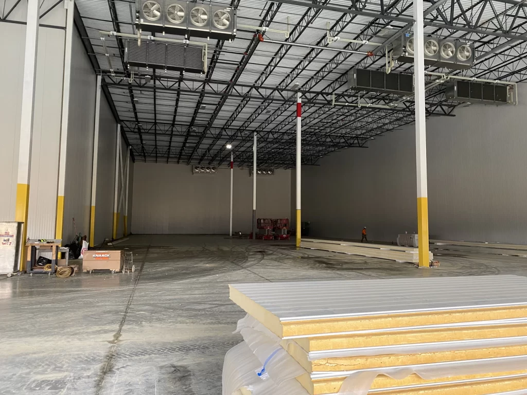 Spacious warehouse interior under construction with metal framework, insulation panels on the floor, and industrial lighting fixtures on the ceiling. Workers and construction materials are visible.