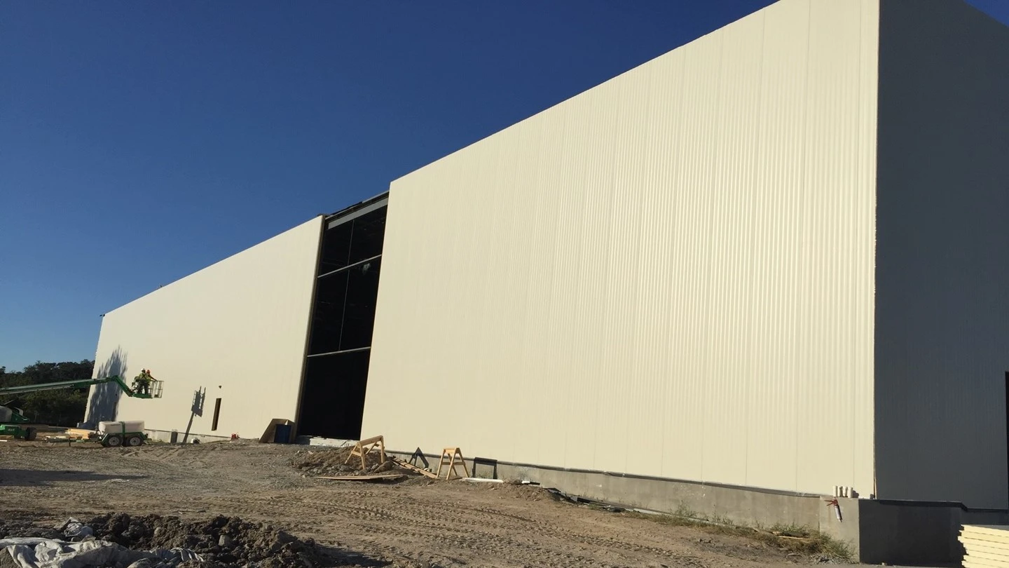 A large, newly constructed industrial building with a partially open side is shown, surrounded by dirt and construction materials under a clear blue sky.