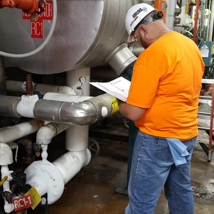 A person wearing a hard hat and an orange shirt is inspecting industrial machinery and taking notes on a clipboard in a facility. Pipes and equipment are visible in the background.