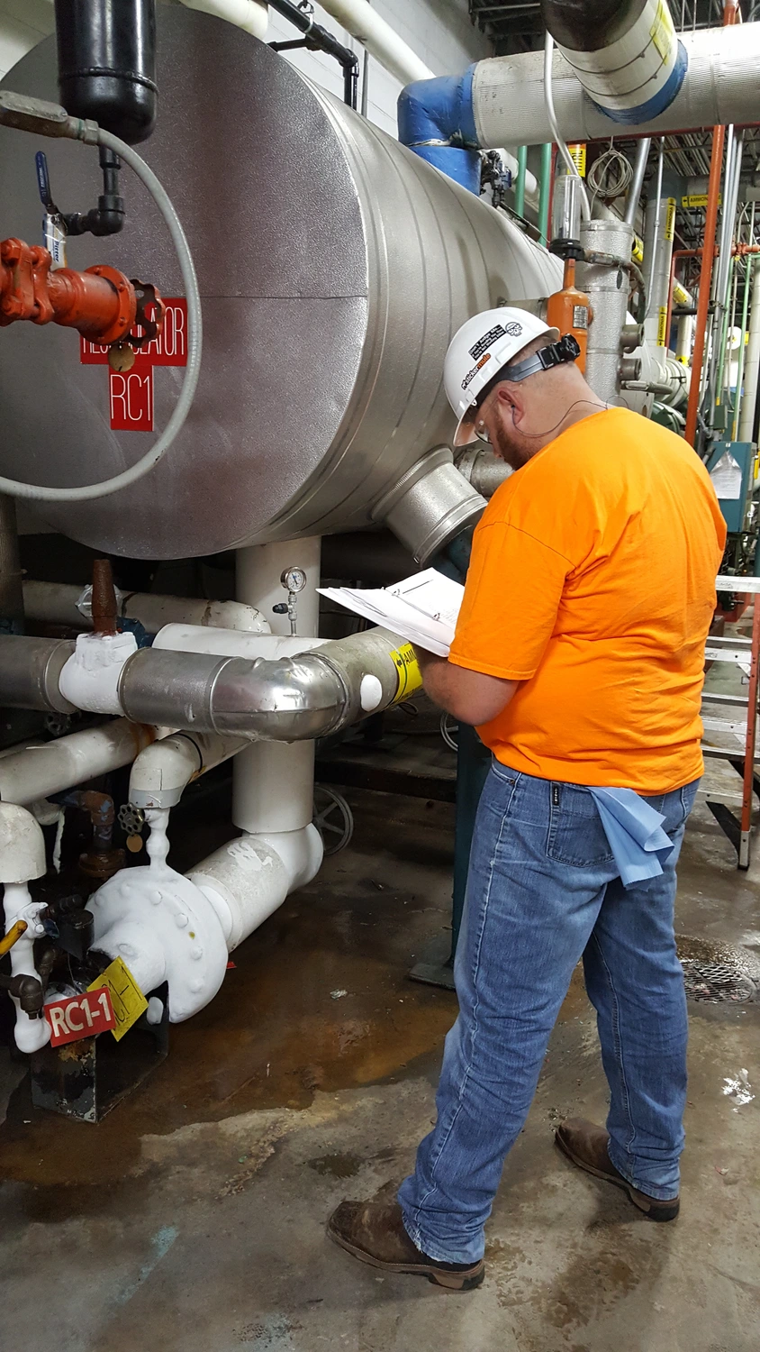 A person wearing a hard hat and an orange shirt is inspecting industrial machinery and taking notes on a clipboard in a facility. Pipes and equipment are visible in the background.